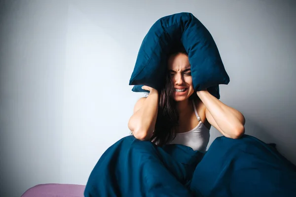 Jonge mooie vrouw in het ochtendbed thuis. Geïrriteerde vrouwelijke persoon lijdt aan lawaai. Houd het kussen rond het hoofd en klaar om te huilen. Alleen in een depressieve kamer. — Stockfoto