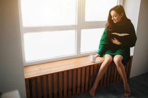 Le matin parfait Une jeune femme brune s'assoit sur le rebord d'une fenêtre et tient dans ses mains un livre et une tasse de thé ou de café. Modèle féminin habillé en pull oversize vert . — Photo