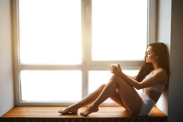 Manhã perfeita Uma jovem morena senta-se em um peitoril da janela e segura uma xícara de chá ou café em suas mãos. Modelo feminino em roupa interior branca — Fotografia de Stock