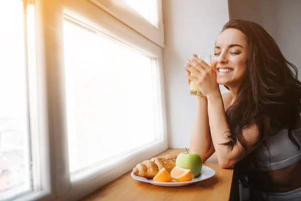 Good morning Young brunette woman in white underwear has breakfast on a wooden windowsill. A young female model eats. There is fresh orange or juice, croissant, fruit, apple on a white plate