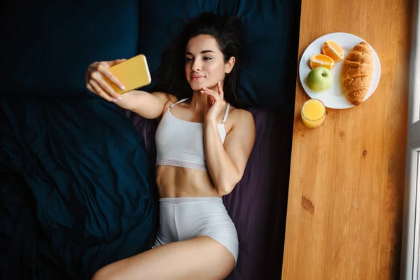 Jeune femme brune en sous-vêtements blancs prend le petit déjeuner sur un rebord de fenêtre en bois. Elle utilise un téléphone. vue de dessus. Il y a de l'orange fraîche ou du jus, croissant, fruit, pomme sur une assiette blanche — Photo