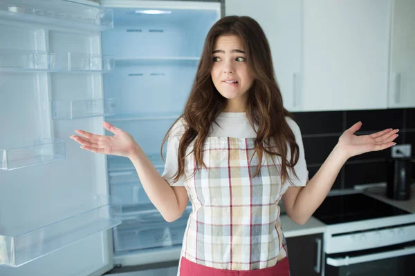 Mujer joven en la cocina durante la cuarentena. Párate frente a la nevera vacía abierta sin frutas ni verduras en ella. No sabe qué hacer o cocinar. . — Foto de Stock