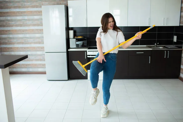 Young woman on kitchen during quarantine. Holding broom in hands as musical instrument and pretend to play on it. Have fun alone after cleaning floor. — Stock Photo, Image
