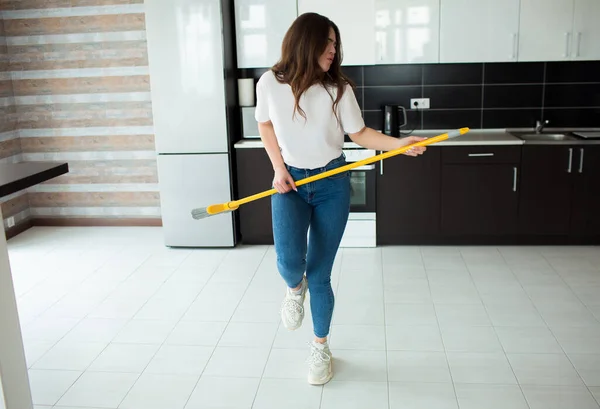 Young woman on kitchen during quarantine. Hold broom in hands as musical instrument and pretend to play on it. Dance alone after cleaning.