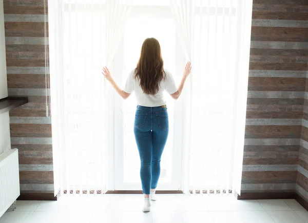 La jeune femme se tient sur la cuisine pendant la quarantaine. Vue arrière de la personne féminine regarder la fenêtre et tenir la main sur les rideaux. Isolement personnel à la maison. Belle lumière du jour blanche . — Photo