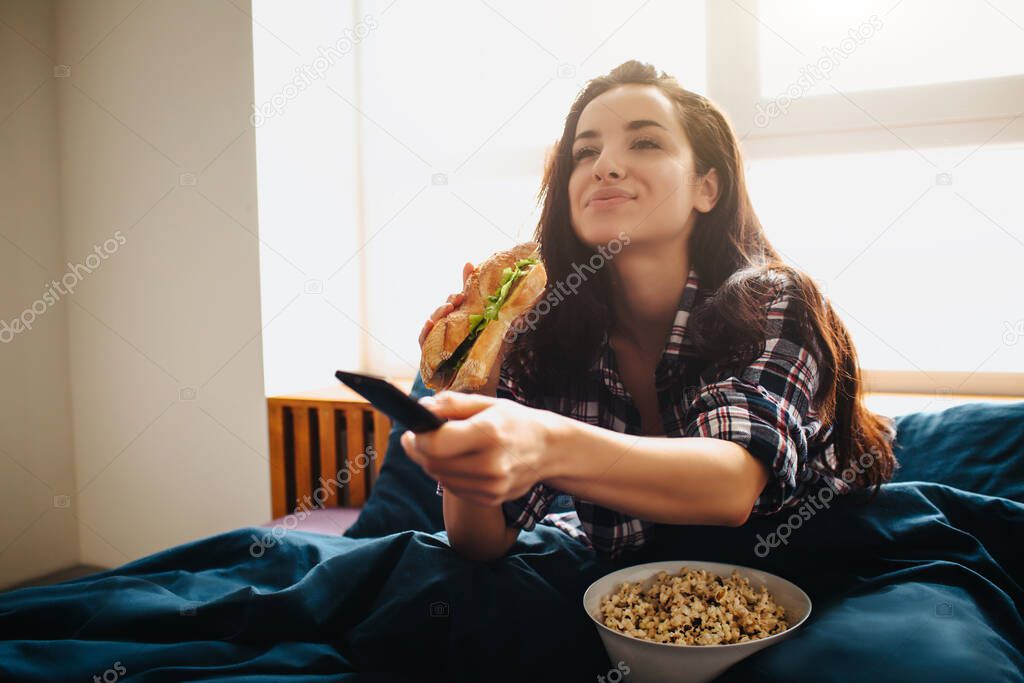 Young beautiful woman in morning bed at home. Excited happy female person watching tv. Using remote control and eating sandwich with popcorn. Movie time.