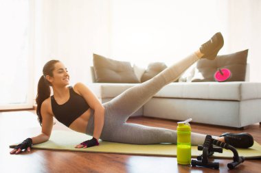 Karantina sırasında spor yapan genç bir kadın. Kız kalça tarafında yatıyor ve sol bacağını yukarıda tutuyor. Vücudun alt kısımlarını esnetiyorum. Odada tek başına egzersiz yapmak..