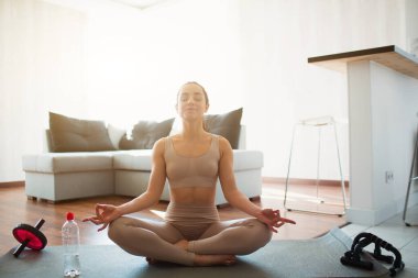 Young woman doing yoga workout in room during quarantine. Sit on mat in lotus position with legs crossed. Meditating alone in room. Water bottle and sports home eqipment besides. clipart
