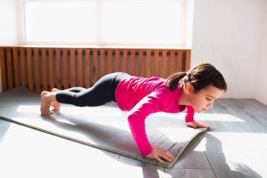 Evde şınav çeken küçük bir kız. Şirin çocuk paspasın üstünde antrenman yapıyor. Koyu renk saçlı kadın model spor giyim bölümünde odasının penceresinin yanında egzersiz yapıyor.