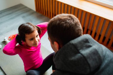 Genç baba ve küçük tatlı kızı evde ABS egzersizi yapıyorlar. Şirin bir çocuk ve babası kapalı alanda paspas eğitimi alıyor. Küçük siyah saçlı kadın model spor giyim bölümünde odanın penceresinin yanında egzersiz yapıyor.