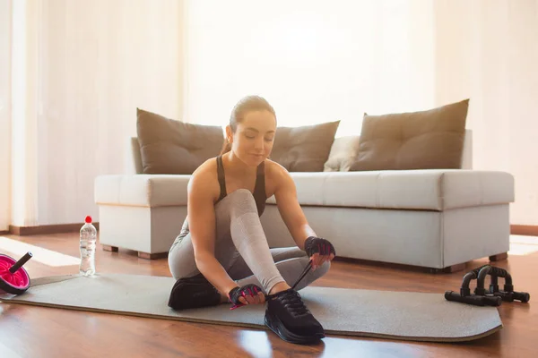 Mujer joven haciendo ejercicio deportivo en la habitación durante la cuarentena. Siéntate en la alfombra y tuing cordones zapatos. Preparación para el entrenamiento en casa . — Foto de Stock