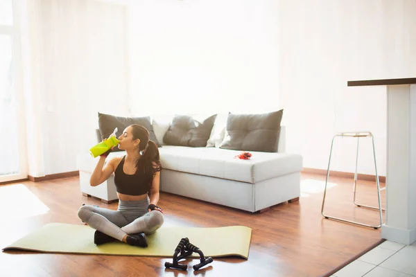 Ung kvinna gör sport träning i rummet under karantän. Sitt på matta med korsade ben och drick protein från grön flaska. Vila koppla av efter träningen. — Stockfoto