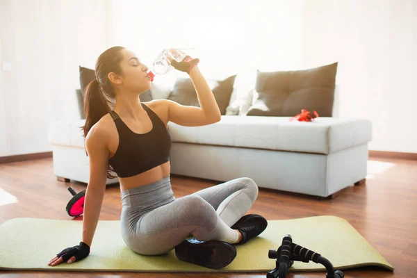 Mujer joven haciendo ejercicio deportivo en la habitación durante la cuarentena. Descanse después del ejercicio. Niña sentarse en la estera y beber agua de la botella de plástico. Pausa después del despertar . — Foto de Stock