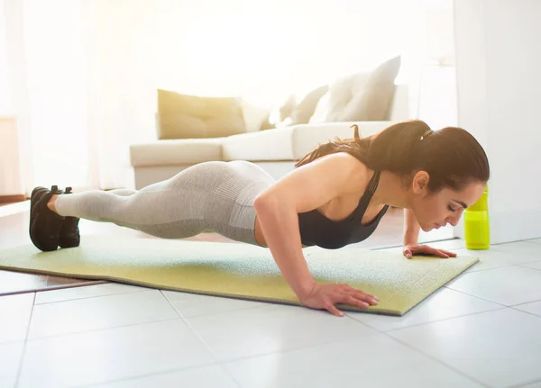 Ung kvinna gör sport träning i rummet under karantän. Stark kraftfull kvinna låg gör push up motion. Stå också i plank position. Träna i rummet på mattan. — Stockfoto