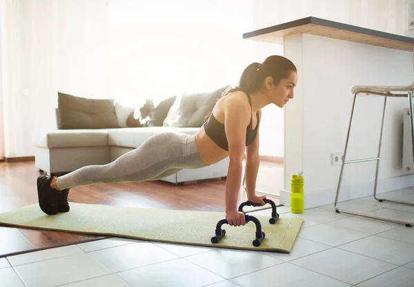 Ung kvinna gör sport träning i rummet under karantän. Bild på ett starkt fitnessmodellställ i plank-läge med hjälp av armhävningshandtag. Motionerar ensam i rummet. — Stockfoto