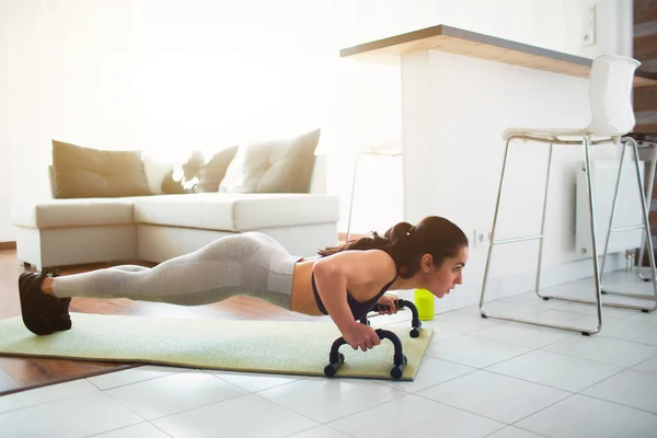 Mujer joven haciendo ejercicio deportivo en la habitación durante la cuarentena. Soporte modelo Fitness en posición de tablón mediante barra de mano push-up. También empujando hacia arriba en la estera . — Foto de Stock
