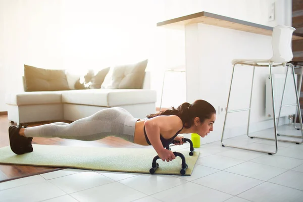 Karantina sırasında spor yapan genç bir kadın. Fitness modeli destek çubuğunu kullanarak tahta pozisyonunda duruyor. Aynı zamanda paspasa basıyorum..