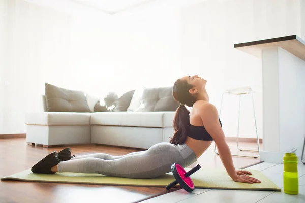 Mujer joven haciendo ejercicio deportivo en la habitación durante la cuarentena. Retrocediendo en la esterilla de yoga. Acostado sobre el estómago y estirándose . — Foto de Stock