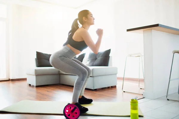Mujer joven haciendo ejercicio deportivo en la habitación durante la cuarentena. Hacer ejercicio en cuclillas en la estera de yoga en la habitación. Entrenamiento concentrado . — Foto de Stock