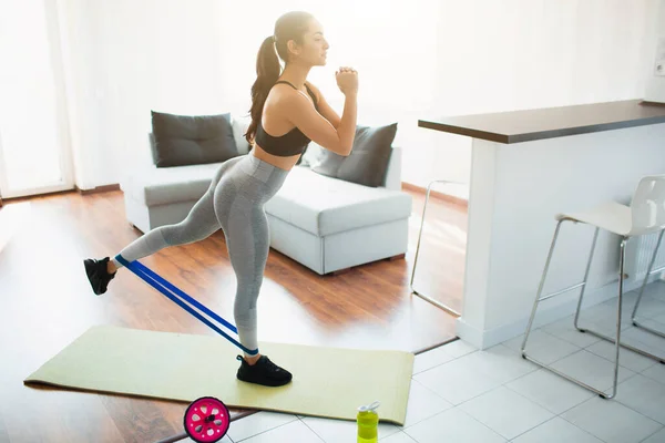 Mujer joven haciendo ejercicio deportivo en la habitación durante la cuarentena. Párese en la esterilla de yoga y estire la pierna en la espalda usando banda de resistencia . — Foto de Stock