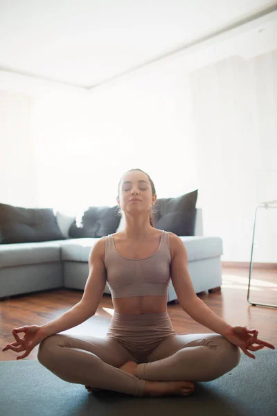 Ung kvinna som gör yoga träning i rummet under karantän. Flickan sitter i lotusposition och mediterar med slutna ögon. Stretching och träning hemma. — Stockfoto