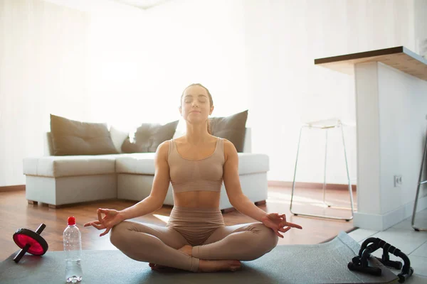 Ung kvinna som gör yoga träning i rummet under karantän. Sitt på mattan i lotusställning med benen korsade. Mediterar ensam i rummet. Vattenflaska och sport hem sändning förutom. — Stockfoto