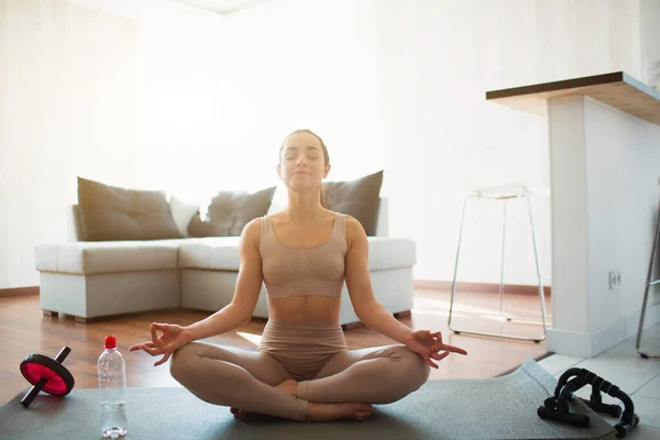 Karantina sırasında odada yoga yapan genç bir kadın. Lotus pozisyonunda bacak bacak üstüne atarak otur. Odada yalnız meditasyon yapmak. Su şişesi ve spor malzemeleri dışında..