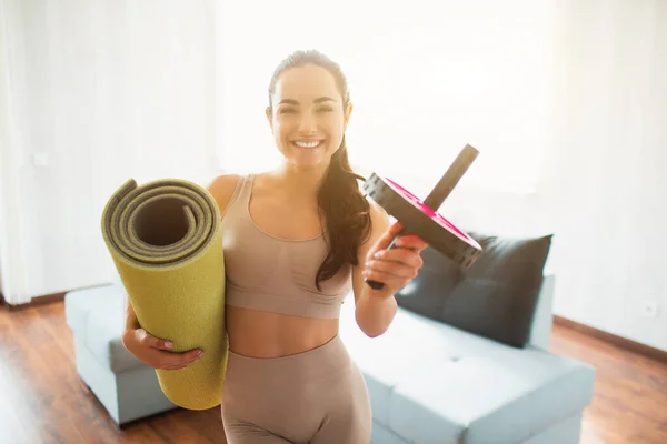 Ung kvinna som gör yoga träning i rummet under karantän. Glada positiva flicka ler och håller rullade yogamatta och buken träningsroller. Efter träning. — Stockfoto