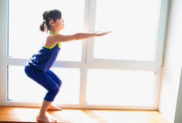 Liten flicka gör squat övningar hemma. Söt unge tränar på ett träfönster inomhus. Lite mörkhårig kvinnlig modell i sportkläder har övningar nära fönstret i sitt rum — Stockfoto