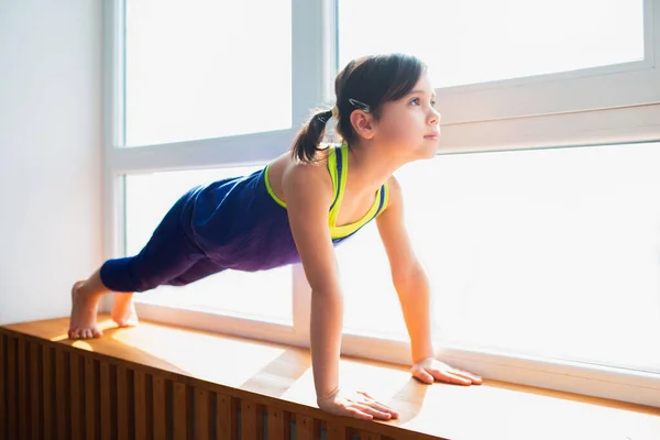Liten flicka gör en Plank Motion träning hemma. Söt unge tränar på ett träfönster inomhus. Lite mörkhårig kvinnlig modell i sportkläder har övningar nära fönstret i sitt rum. — Stockfoto