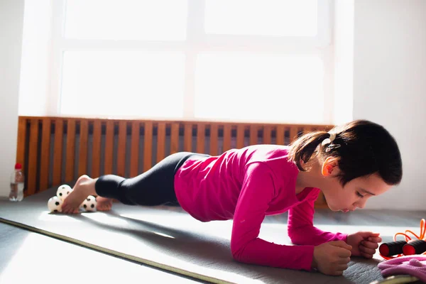 Liten flicka gör en Plank Motion träning hemma. Söt unge tränar på en matta inomhus. Lite mörkhårig kvinnlig modell i sportkläder har övningar nära fönstret i sitt rum — Stockfoto