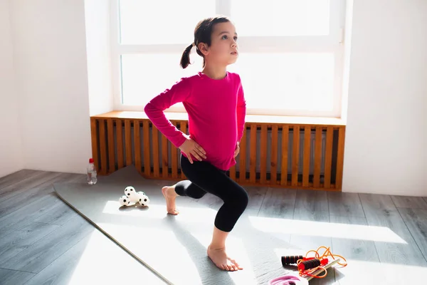 Liten flicka framåt lunge träning hemma. Söt unge tränar på en matta inomhus. Lite mörkhårig kvinnlig modell i sportkläder har övningar nära fönstret i sitt rum — Stockfoto