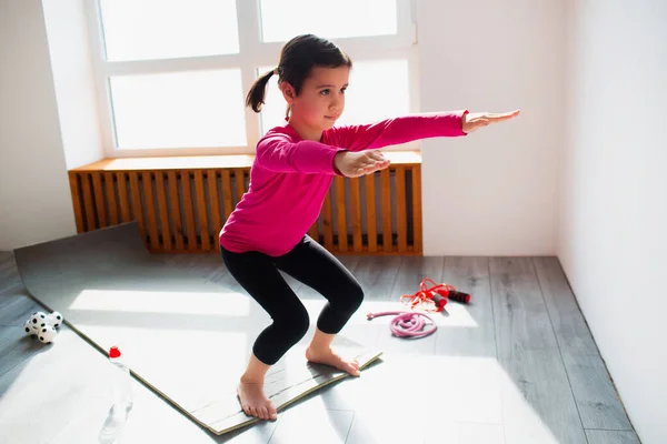 Holčička doma cvičí dřepy. Roztomilé dítě trénuje na rohožce uvnitř. Malá tmavovlasá modelka ve sportovním oblečení má cvičení u okna ve svém pokoji — Stock fotografie