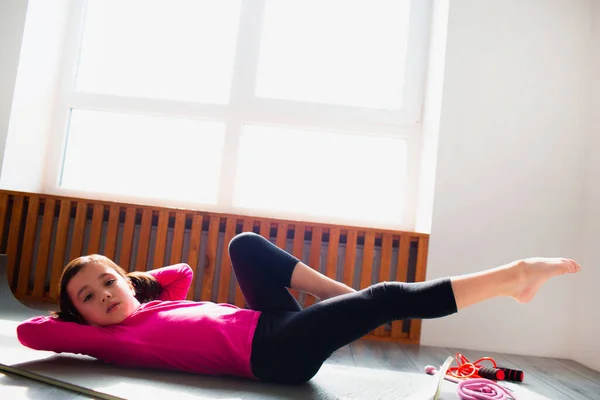 Liten flicka gör ABS träning hemma. Söt unge tränar på en matta inomhus. Lite mörkhårig kvinnlig modell i sportkläder har övningar nära fönstret i sitt rum — Stockfoto