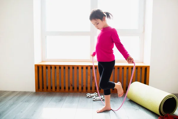 Liten flicka hoppar på ett rep träning hemma. Söt unge tränar på en matta inomhus. Lite mörkhårig kvinnlig modell i sportkläder har övningar nära fönstret i sitt rum — Stockfoto