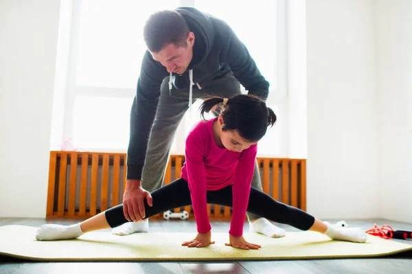 Liten flicka gör stretching övningar träning hemma. Söta barn och pappa tränar på en matta inomhus. Lite mörkhårig kvinnlig modell i sportkläder har övningar nära fönstret i sitt rum — Stockfoto