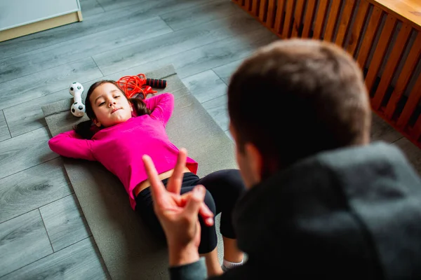 Ung far och hans söta lilla dotter har ABS träning hemma. Söta barn och pappa tränar på en matta inomhus. Lite mörkhårig kvinnlig modell i sportkläder har övningar nära fönstret i rummet — Stockfoto