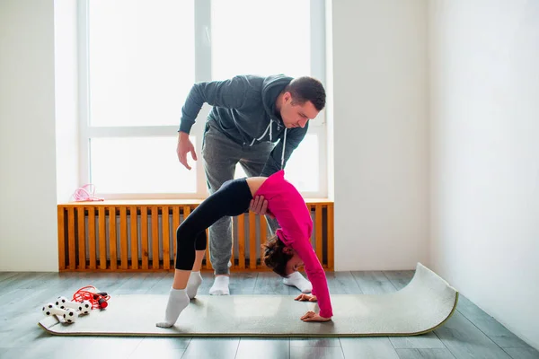 Ung far och hans söta lilla dotter har träning hemma i bro pose. Söta barn och pappa tränar på en matta inomhus. De bär sportkläder och har övningar nära fönstret i rummet — Stockfoto
