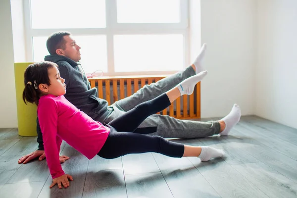 Ung far och hans söta lilla dotter gör reverce planka med ben höja på golvet hemma. Familjens konditionsträning. Söta barn och pappa tränar på en matta inomhus nära fönstret i rummet. — Stockfoto