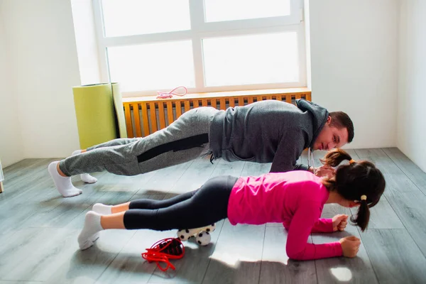 young father and his cute little daughter are doing plank on the floor at home. Family fitness workout. Cute kid and daddy is training on a mat indoor and have exercises near the window in room.