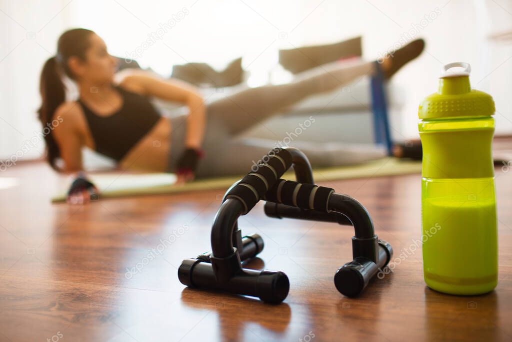 Young woman doing sport workout in room during quarantine. Green protein bottle and push ups hand bar in front. Girl exercising using resistance band. Stretch left leg up and forward.
