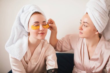 Gözlerinin altında yamalarla eğlenen iki genç kadın. Havlu ve pijamalı iki arkadaş evde eğlenceli bir spa partisi veriyor..