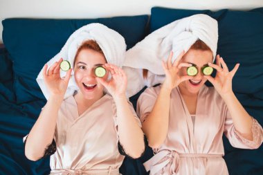 İki kadın model taze yeşil salatalık kullanarak bir göz maskesi yapıyor. Havlu ve pijamalı iki genç kadın evde eğlenceli bir spa partisi veriyor..
