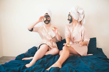 İki kadın model yüz maskesi yapıyor ve taze yeşil salatalık kullanıyor. Havlu ve pijamalı iki genç kadın evde eğlenceli bir spa partisi veriyor..