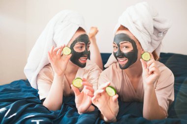 İki kadın model yüz maskesi yapıyor ve taze yeşil salatalık kullanıyor. Havlu ve pijamalı iki genç kadın evde eğlenceli bir spa partisi veriyor..