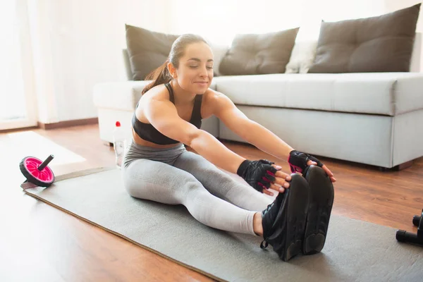 Ung kvinna gör sport träning i rummet under karantän. Sitter på mattan och sträcker sig till fötter eller tår. Uppvärmning före träning. — Stockfoto