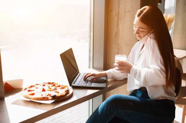 Modelo feminino funciona no laptop e fala ao telefone. Jovem bebe café em um café e fazer um acordo . — Fotografia de Stock