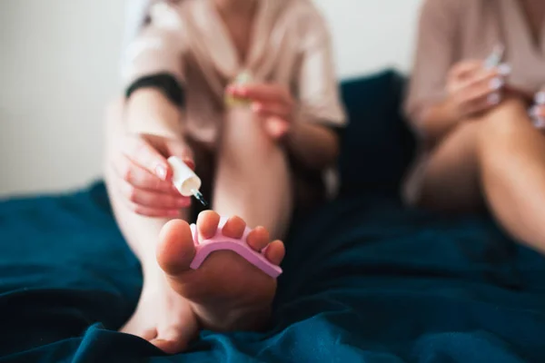 Two young women in towels and pajamas have a fun spa party together in home. They are sitting on the bed and making themselves a new pedicure