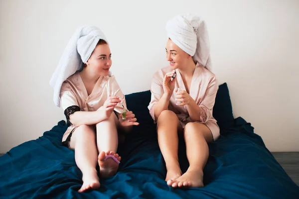 Due giovani donne in asciugamani e pigiama hanno una divertente festa termale insieme a casa. Sono seduti sul letto e si stanno facendo una nuova pedicure — Foto Stock