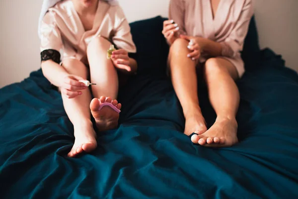 Dvě mladé ženy v ručnících a pyžamu si doma užívají zábavnou lázeňskou párty. Sedí na posteli a dělají si novou pedikúru. — Stock fotografie